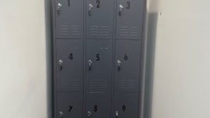 Graue Metallschließfächer mit den Nummern 1 bis 9, angeordnet in zwei Säulen, in einem schmalen Flur mit Holzboden und weißen Wänden.
