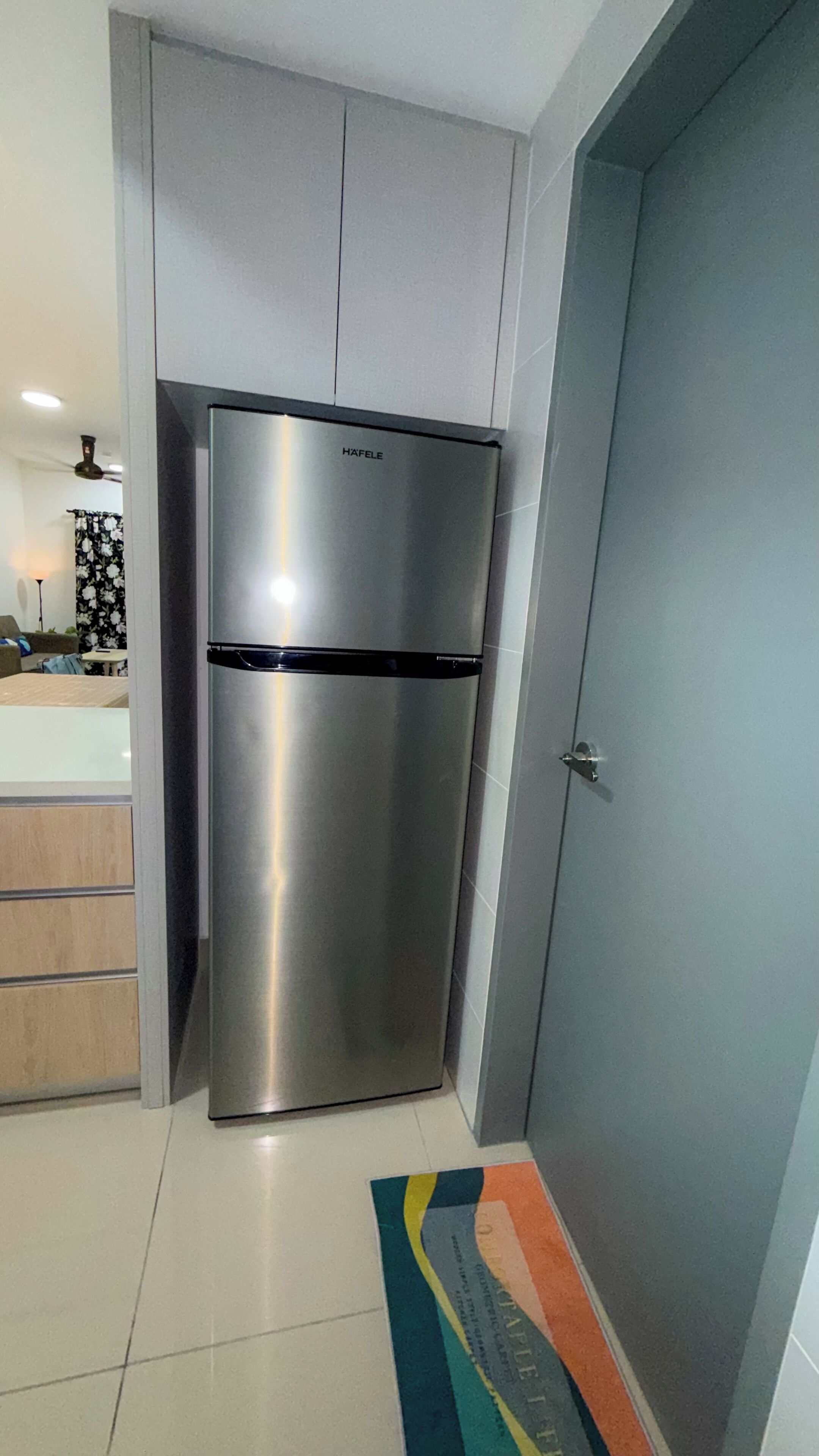 A modern kitchen with a stainless steel refrigerator, a grey door, and a tiled floor.