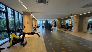 Academia com equipamentos de ginástica, janelas amplas e piso de madeira.
