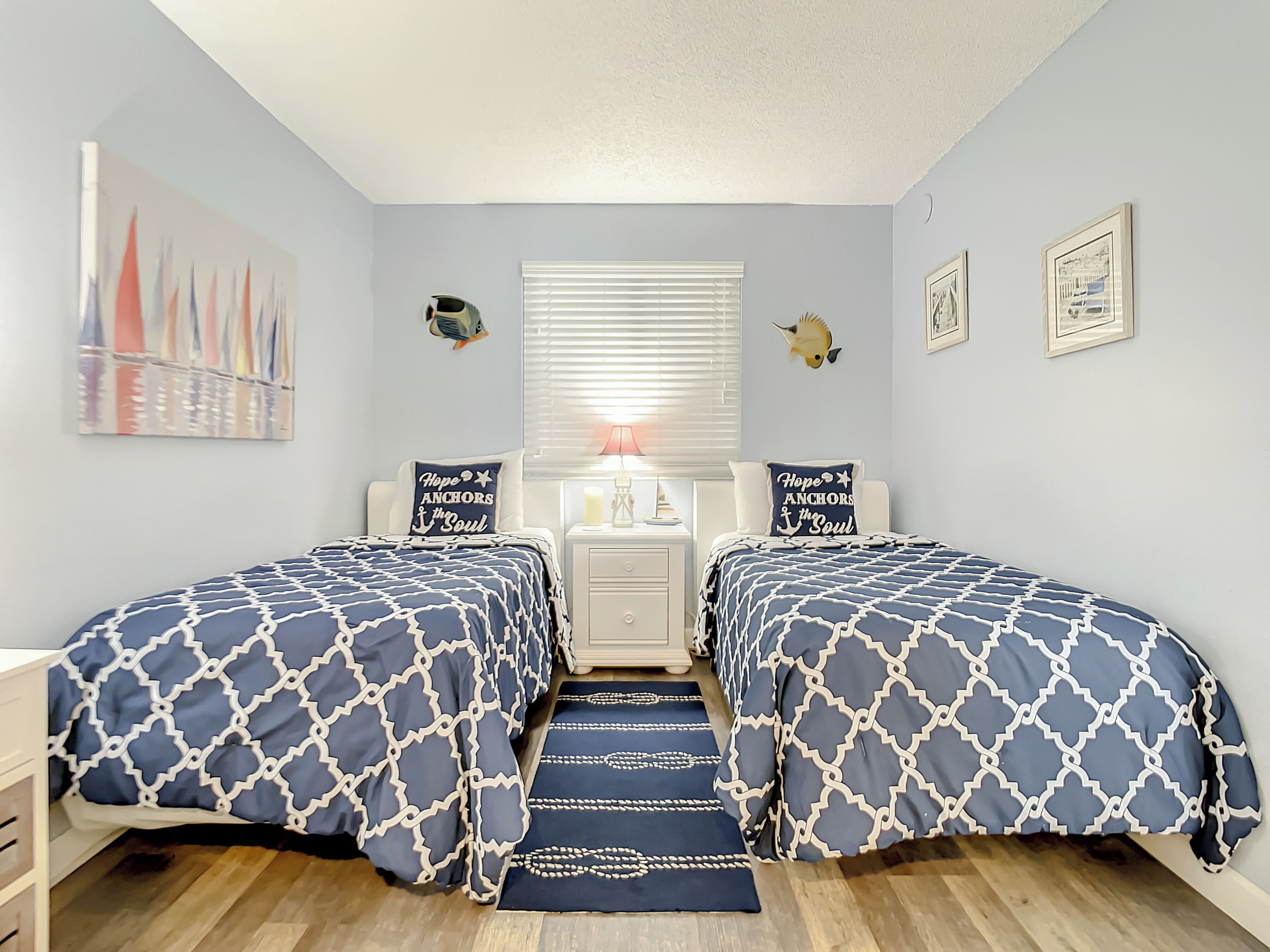 A bedroom with two beds, a nightstand, a lamp, and wall decorations including a fish and a sailboat painting.