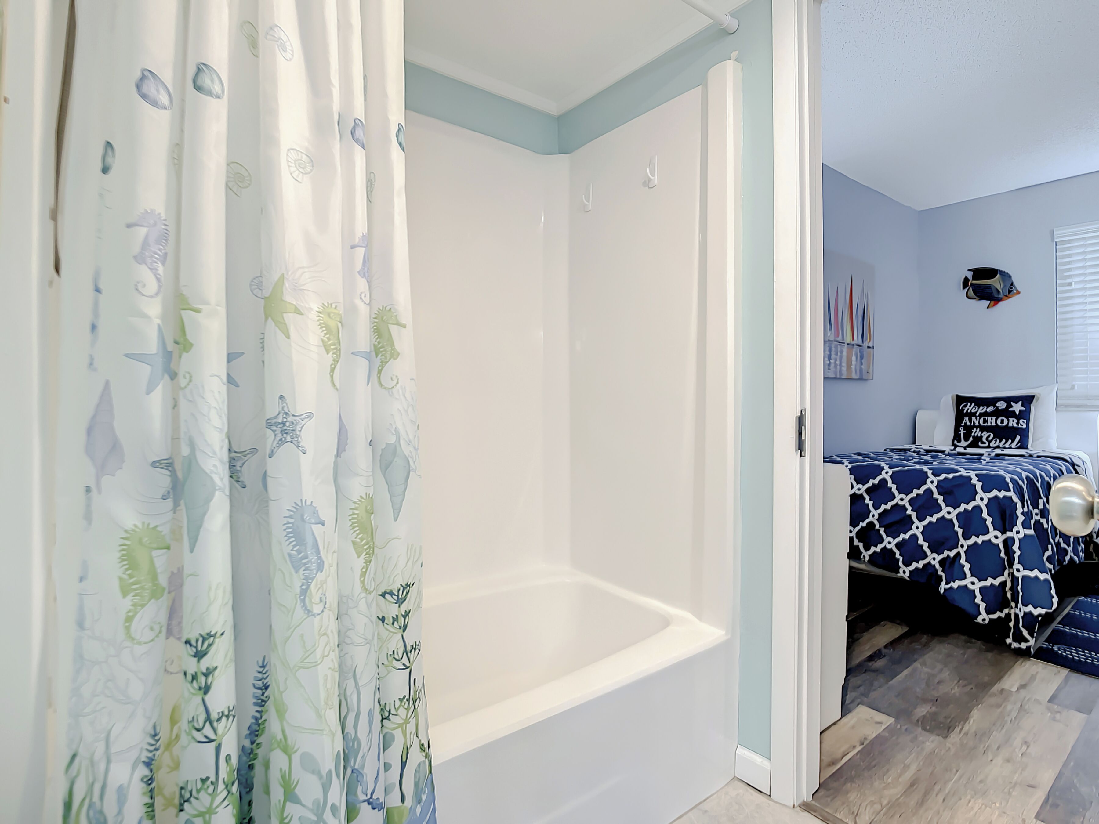 A bathroom with a bathtub, a shower curtain with sea creature patterns, and a bed with a nautical-themed bedspread.
