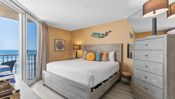 A bedroom with a large bed, a dresser, a nightstand, and a view of the ocean. - Oceania Beach Club 604 (New Smyrna Beach)