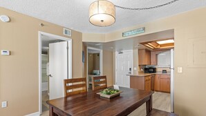 A dining area with a wooden table and chairs, a small side table with a plant, and a kitchen area with wooden cabinets and a microwave. - Oceania Beach Club 604 (New Smyrna Beach)