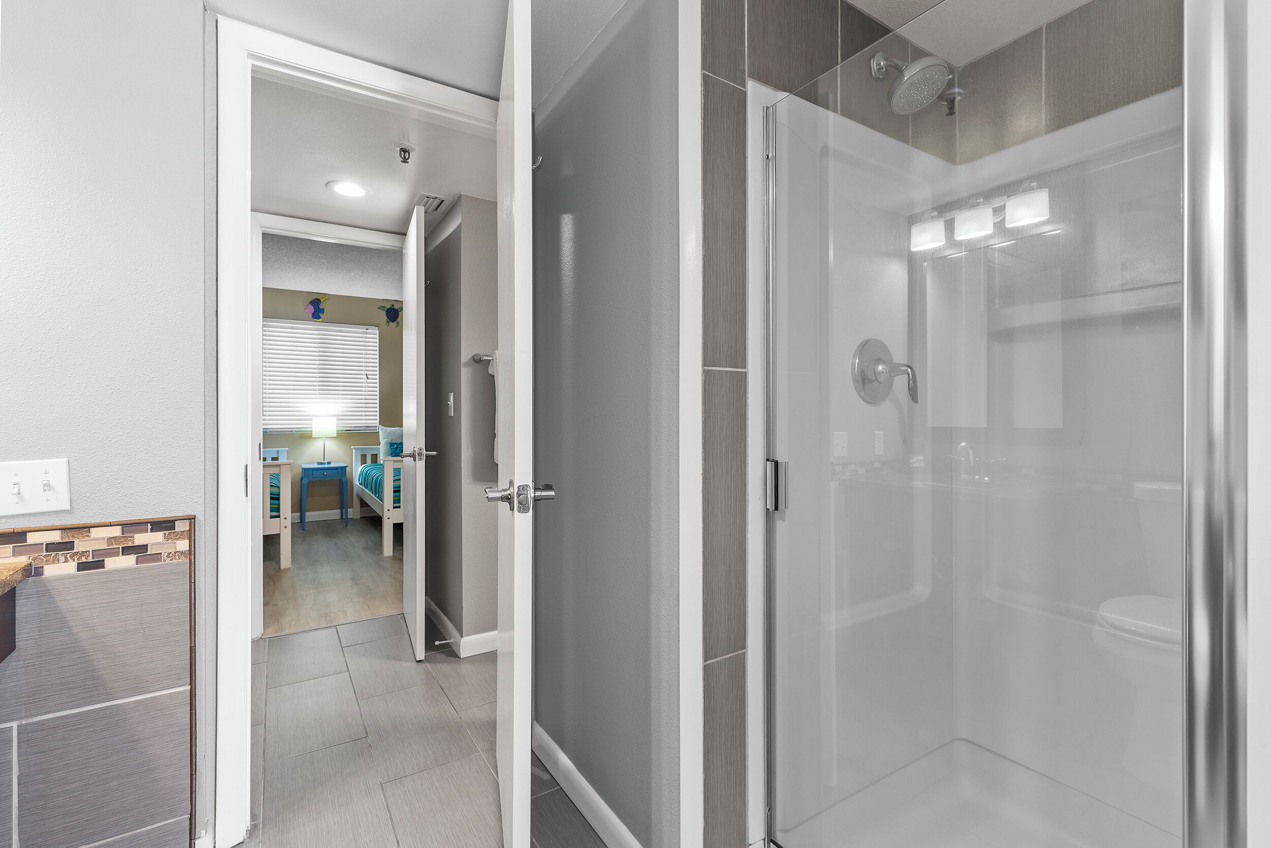 Ein modernes Badezimmer mit Duschkabine aus Glas, sichtbarer Toilette und einer Tür zu einem Raum mit Bett und Schreibtisch.