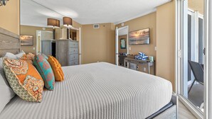 A bedroom with a bed, pillows, a dresser, a desk, and a sliding glass door to a balcony. - Oceania Beach Club 604 (New Smyrna Beach)