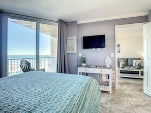 Ein Schlafzimmer mit einem großen Bett, einem Fernseher, einem Schreibtisch mit Pflanze und Blick auf den Strand.