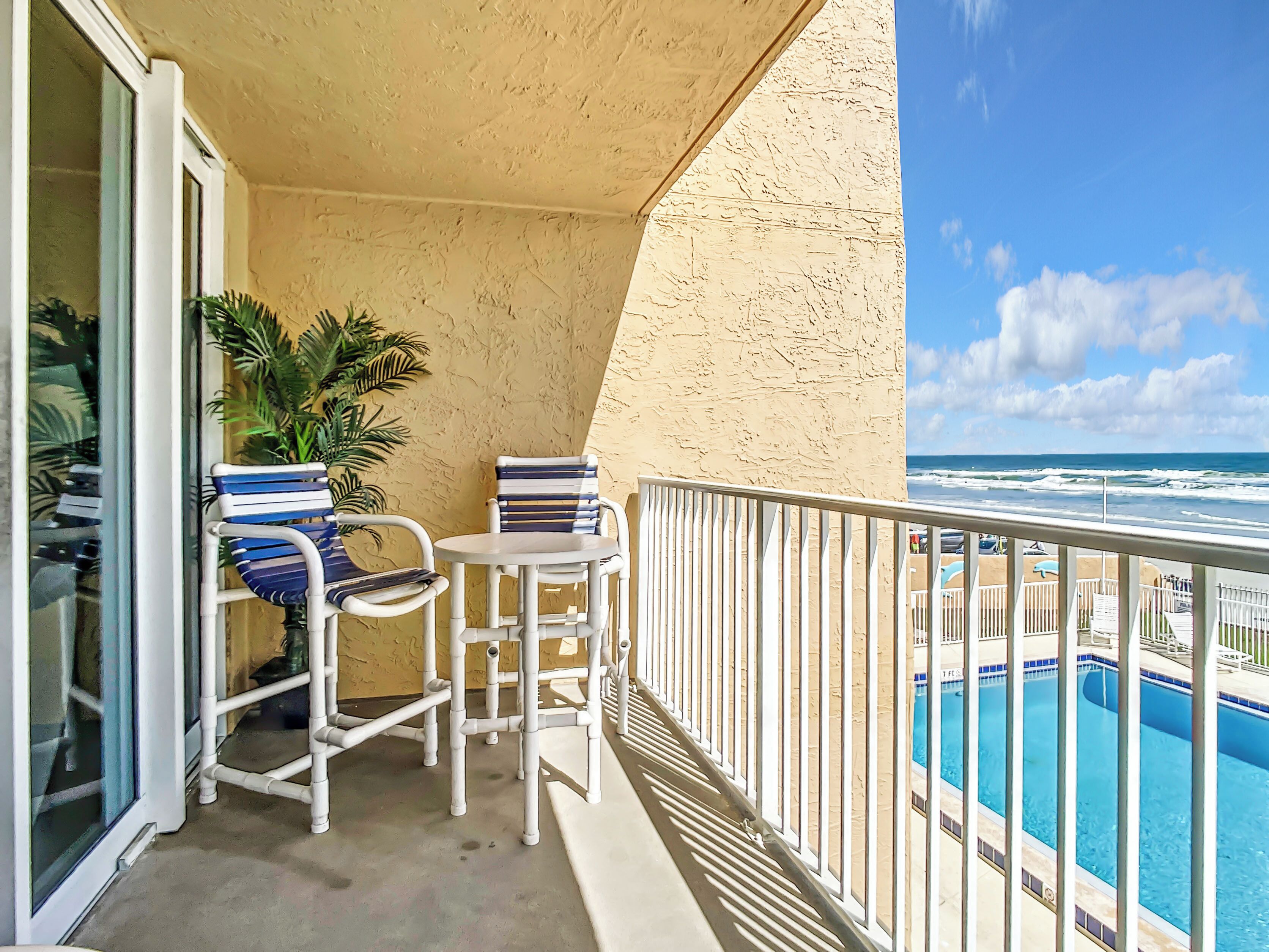 A balcony with two chairs and a small table, overlooking a pool and the ocean.