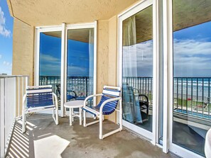 A balcony with two chairs and a small table, overlooking a beach with a railing. - Oceania Beach Club 205 (New Smyrna Beach)