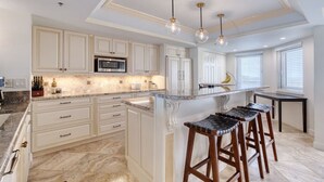 A kitchen with white cabinetry, a central island with stools, a microwave, and pendant lights. - New Smyrna Beachfront Modern 9th Floor Condo Pool Hot Tub (New Smyrna Beach)