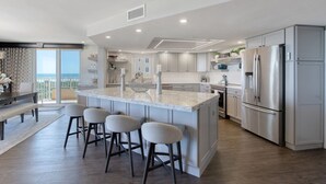 Une cuisine moderne dotée d’un grand îlot central, d’appareils électroménagers en acier inoxydable et offrant une vue sur l’extérieur.