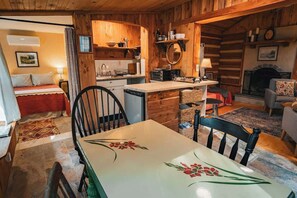 A cozy log cabin interior with a dining table, chairs, a kitchen area, a fireplace, and a bed. - Hawthorne Cabin ~ NEW ~ Romantic ~ Unique ~ Serene (Nellysford)