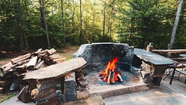 A stone fire pit surrounded by a stone bench and a table, set in a forested area. - Hawthorne Cabin ~ NEW ~ Romantic ~ Unique ~ Serene (Nellysford)