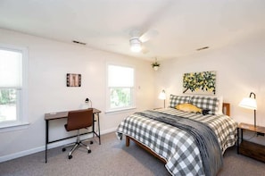 Une chambre à coucher comprenant un lit, un bureau, une chaise, deux fenêtres, un ventilateur de plafond et une horloge murale.