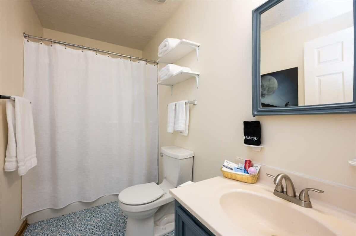 Baño con cortina de ducha, inodoro, lavamanos y espejo que refleja una puerta.