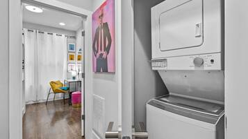 Une buanderie moderne avec un grand sèche-linge industriel et un lave-linge fixé au mur. On aperçoit, à l'arrière-plan, une chaise jaune vif et un pouf rose.