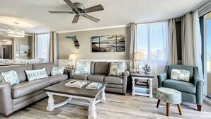 A spacious living room with a ceiling fan, a sectional sofa, a coffee table, and a side table. There are two windows with curtains, a painting on the wall, and a view of the outside. - Oceania Beach Club 106 (New Smyrna Beach)