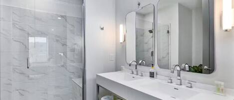 Un baño moderno con cabina de ducha de vidrio, tocador blanco con dos lavamanos y un espejo grande.