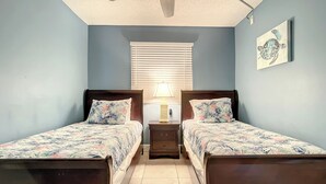 A bedroom with two single beds, a ceiling fan, a window with blinds, a bedside table with a lamp, and a wall-mounted picture. - Oceania Beach Club 606 (New Smyrna Beach)