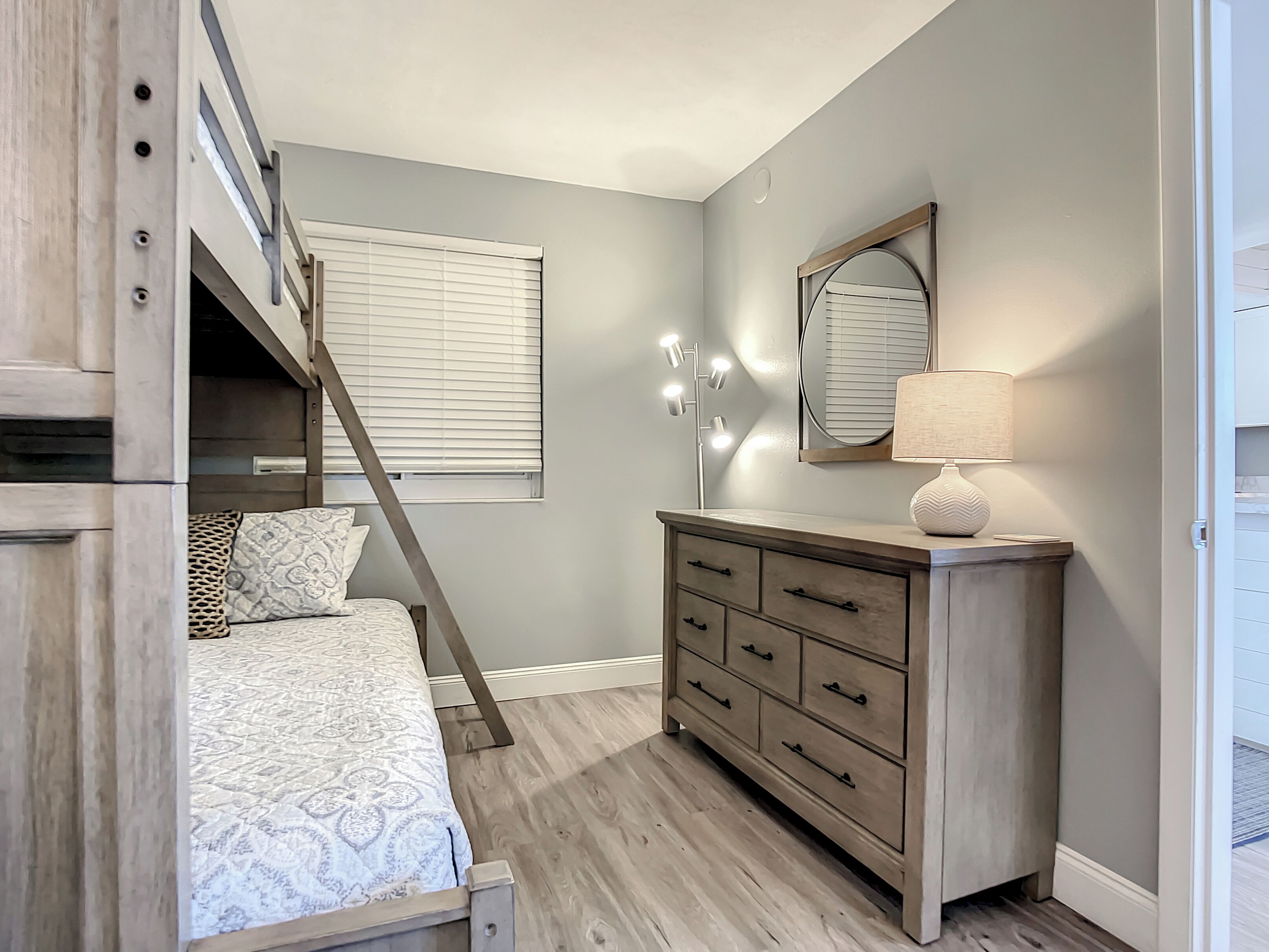 A bedroom with a bunk bed, a dresser, a mirror with lights, and a window with blinds.