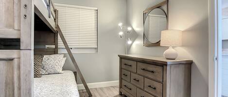 A bedroom with a bunk bed, a dresser, a mirror with lights, and a window with blinds.