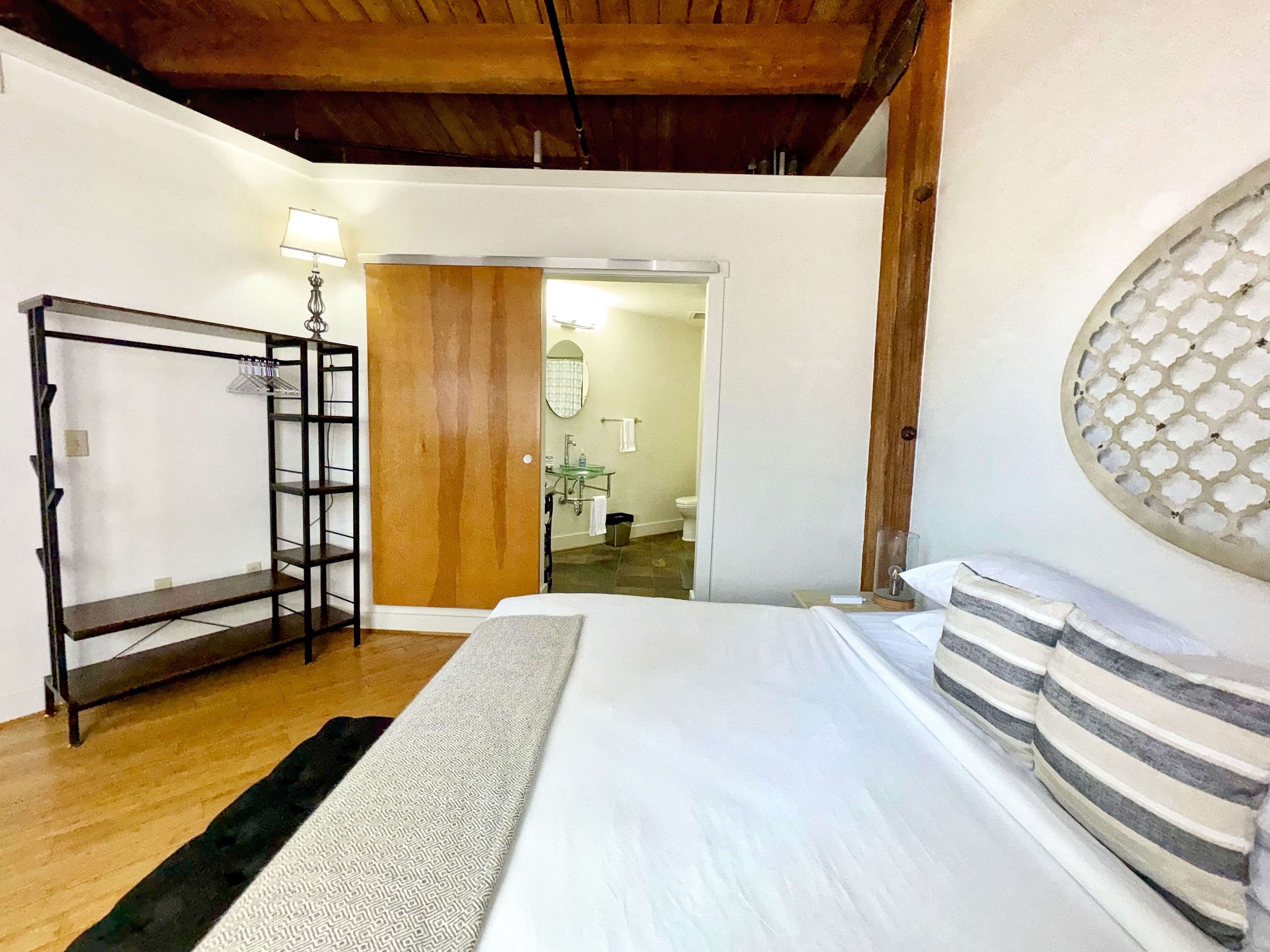 Ein Schlafzimmer mit einem Bett, einem Holzschrank, einem Spiegel und einem durch eine offene Tür einsehbaren Badezimmer.