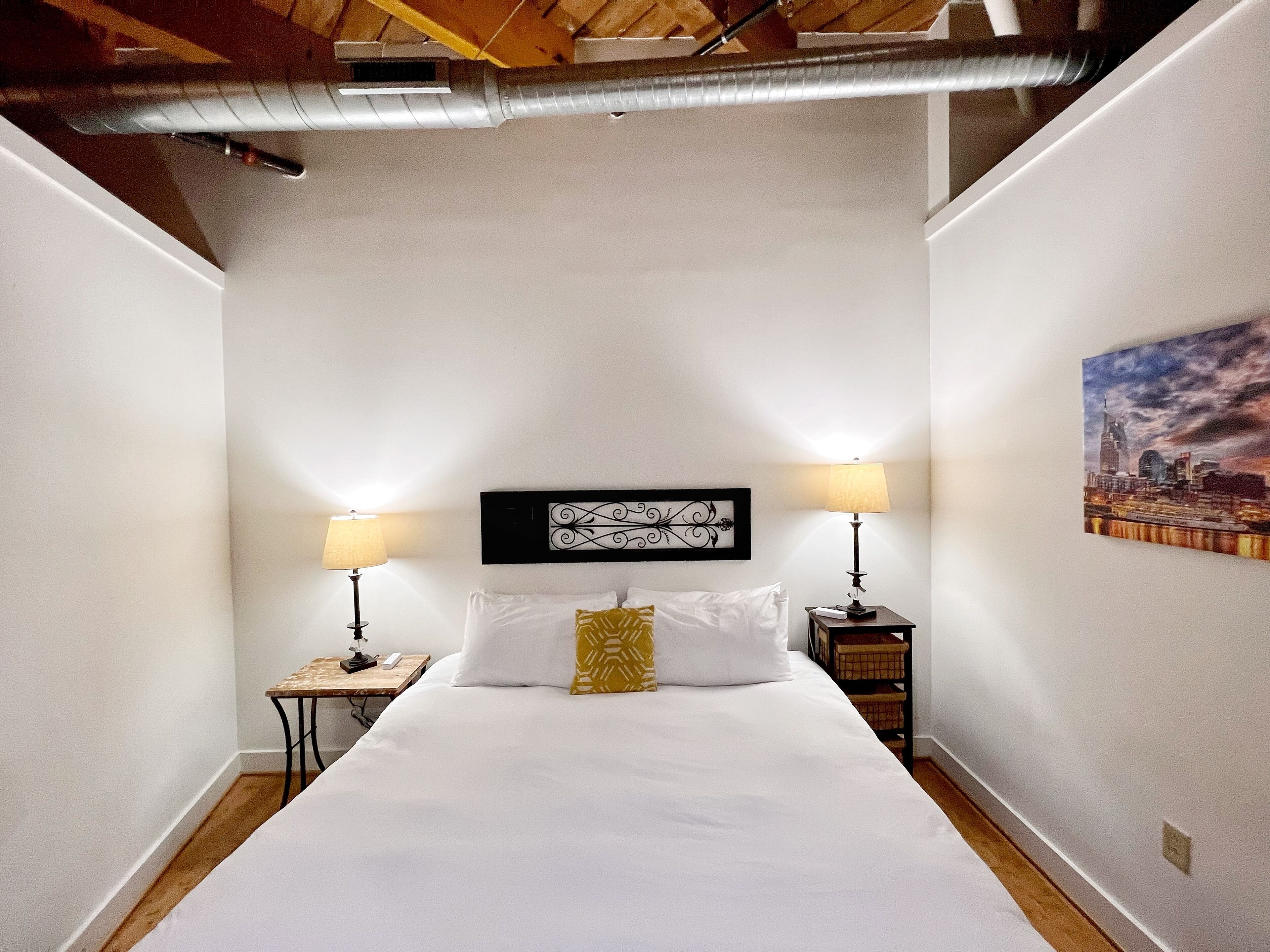 Una habitación de hotel con una cama grande, dos mesitas de noche con lámparas, un adorno en la pared sobre la cama y un cuadro de un paisaje urbano.