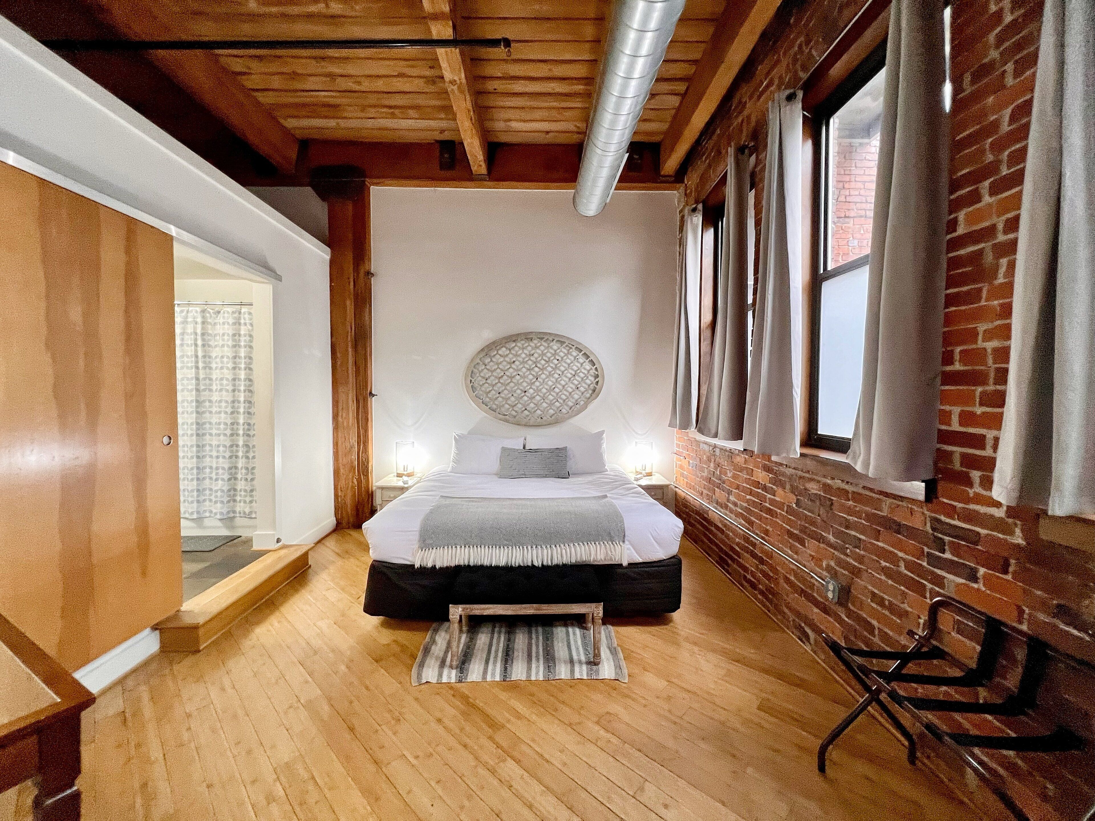 A bedroom with a bed, a round wall-mounted light fixture, a window with curtains, and a brick wall.