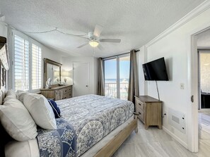 A bedroom with a bed, a ceiling fan, a TV, and a window with a view of the beach. - Oceania Beach Club 409 (New Smyrna Beach)