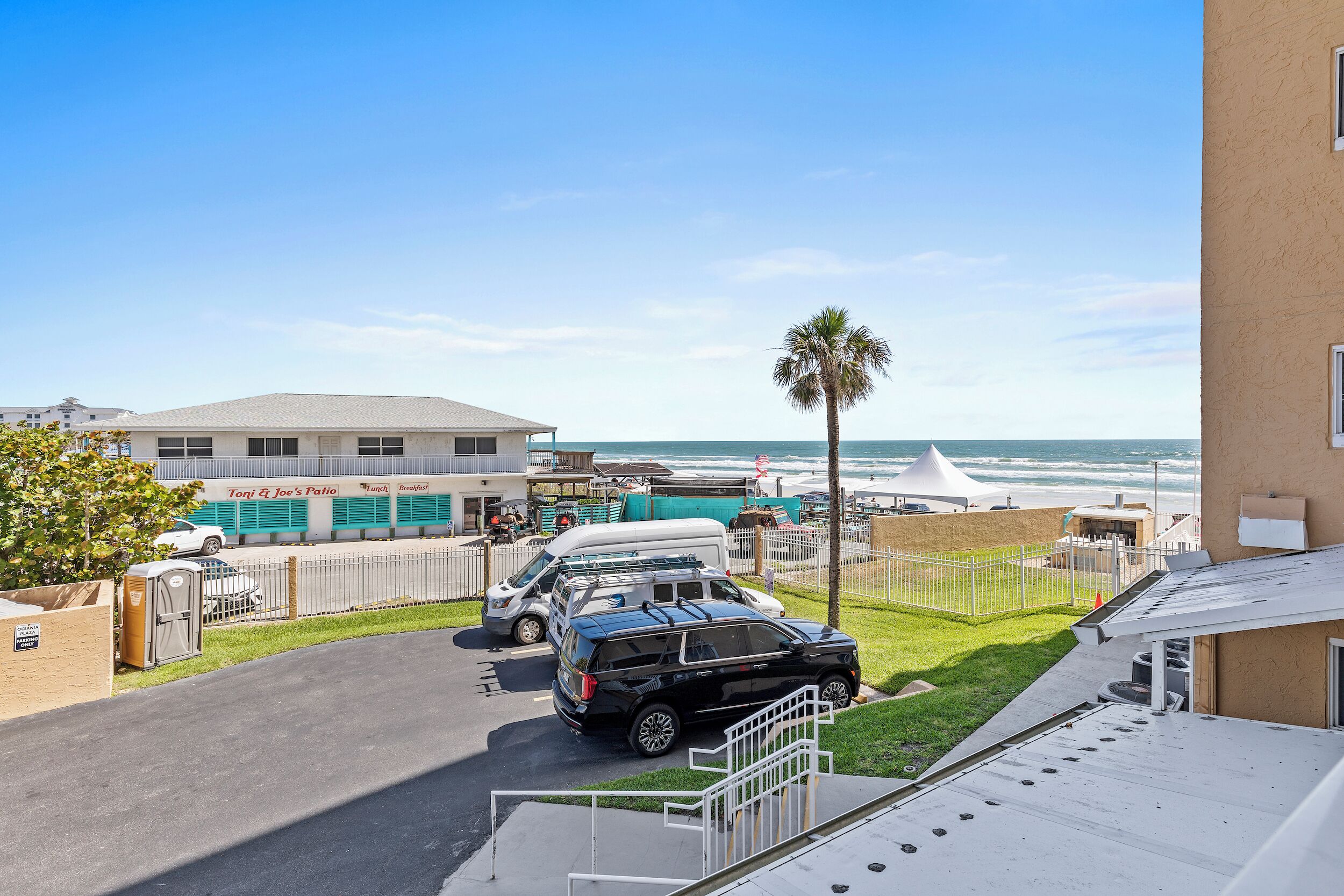 Um estacionamento com um SUV preto, uma van branca e um carro branco. Há um edifício com uma placa que diz “Tony & Joe's Patio”. Uma palmeira é visível e o oceano ao fundo.