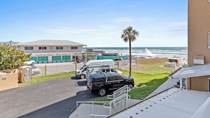 A parking area with a black SUV, a white van, and a white car. There is a building with a sign that reads "Tony & Joe's Patio." A palm tree is visible, and the ocean is in the background. - Oceania Beach Club 207 (New Smyrna Beach)