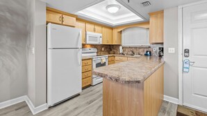 A modern kitchen with wooden cabinets, a white refrigerator, a microwave, a stove, and a granite countertop island. - Oceania Beach Club 207 (New Smyrna Beach)