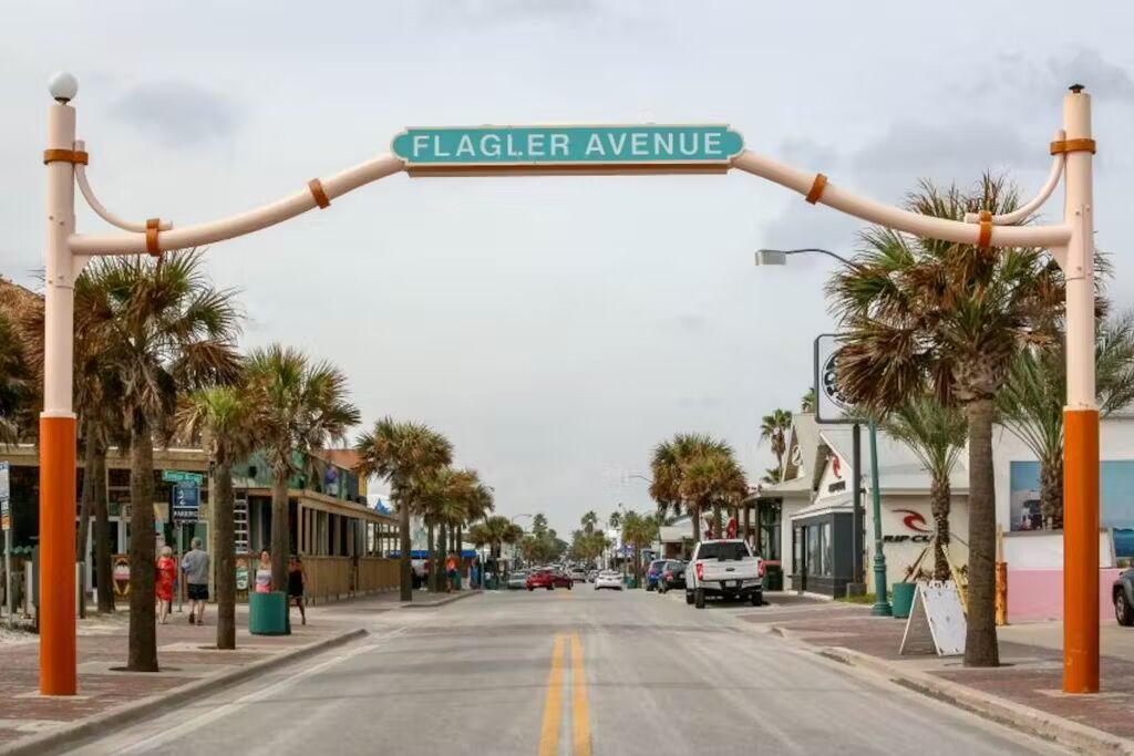A street view of Flagler Avenue with palm trees lining the road, storefronts on both sides, and a clear sky.
