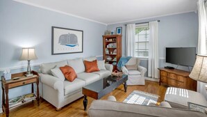 A living room with a sofa, armchair, coffee table, bookshelf, and a television. - Neighborhood Retreat - Large Yard Family Fire Pit (Nashville)