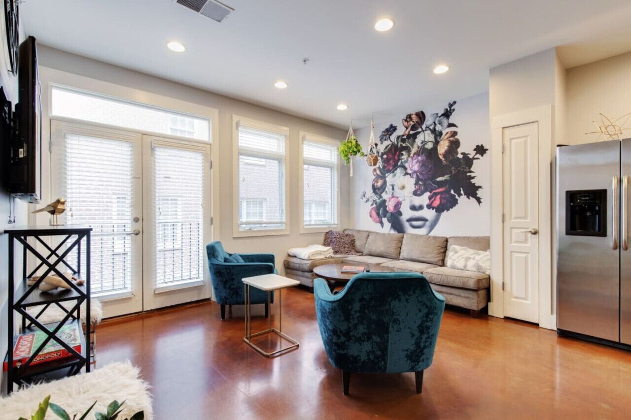 A modern living room with a sofa, two armchairs, a coffee table, and a TV. There are large windows with blinds, a refrigerator, and a decorative wall piece.