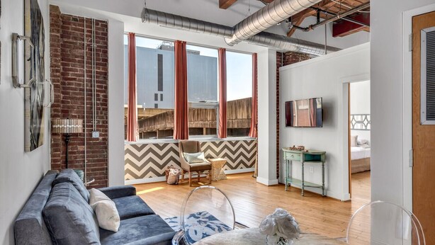 A modern living room with a sofa, a coffee table, and a TV mounted on the wall. There is a brick wall and a window with curtains. - June (Nashville)