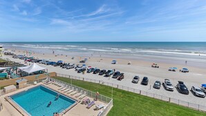 A beach view with a swimming pool, beach umbrellas, and parked cars. - Oceania Beach Club 503 (New Smyrna Beach)