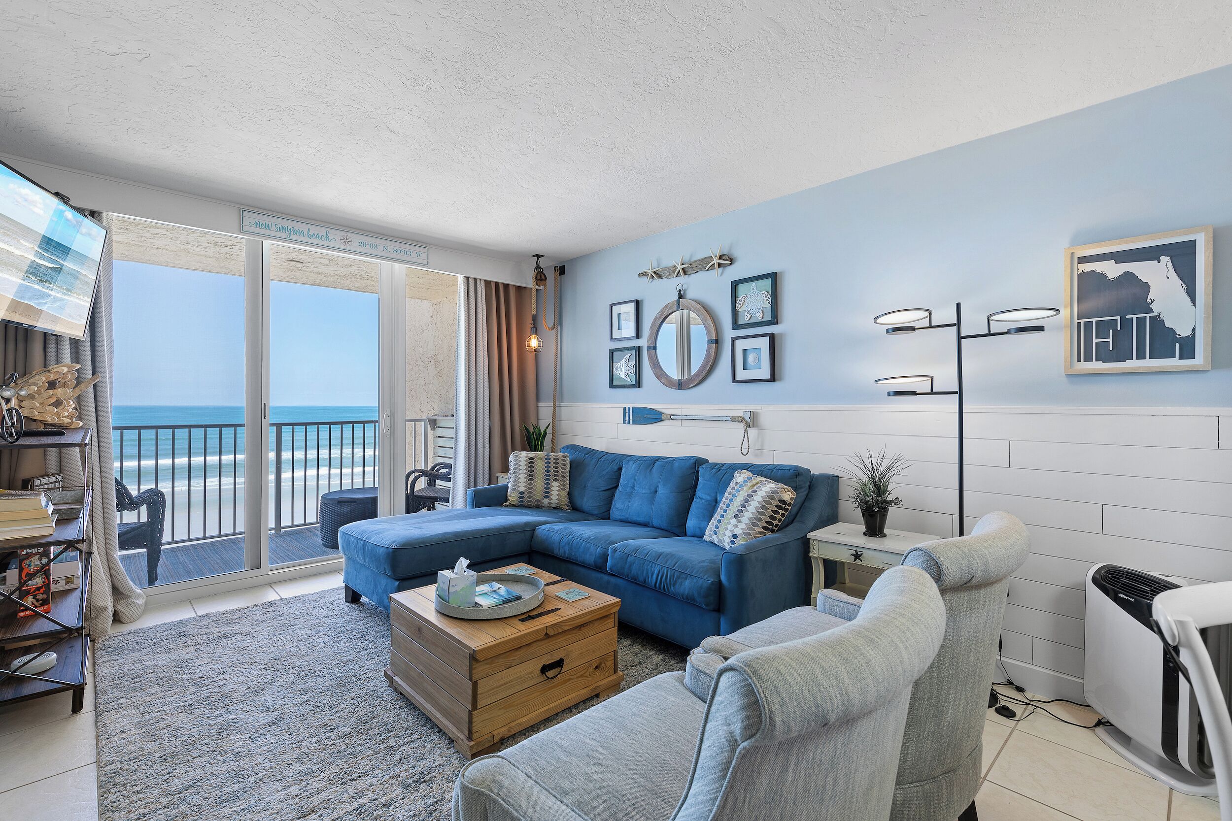 A living room with a blue sofa, a wooden coffee table, and a grey armchair. There is a large window with a view of the ocean.