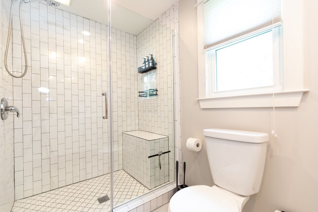 Ein modernes Badezimmer mit weißen Fliesen, einer gläsernen Duschkabine, einer Toilette und einem Fenster für natürliches Licht.