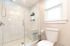 Et moderne badeværelse med hvide flisemure, et glasafskærmet brusebad, toilet og et vindue med naturligt lys.