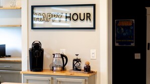 Un comptoir de cuisine avec une machine à café, une bouilloire et une cafetière à piston. Un panneau indiquant « HEURE HEUREUSE » est affiché sur le mur au-dessus du comptoir.