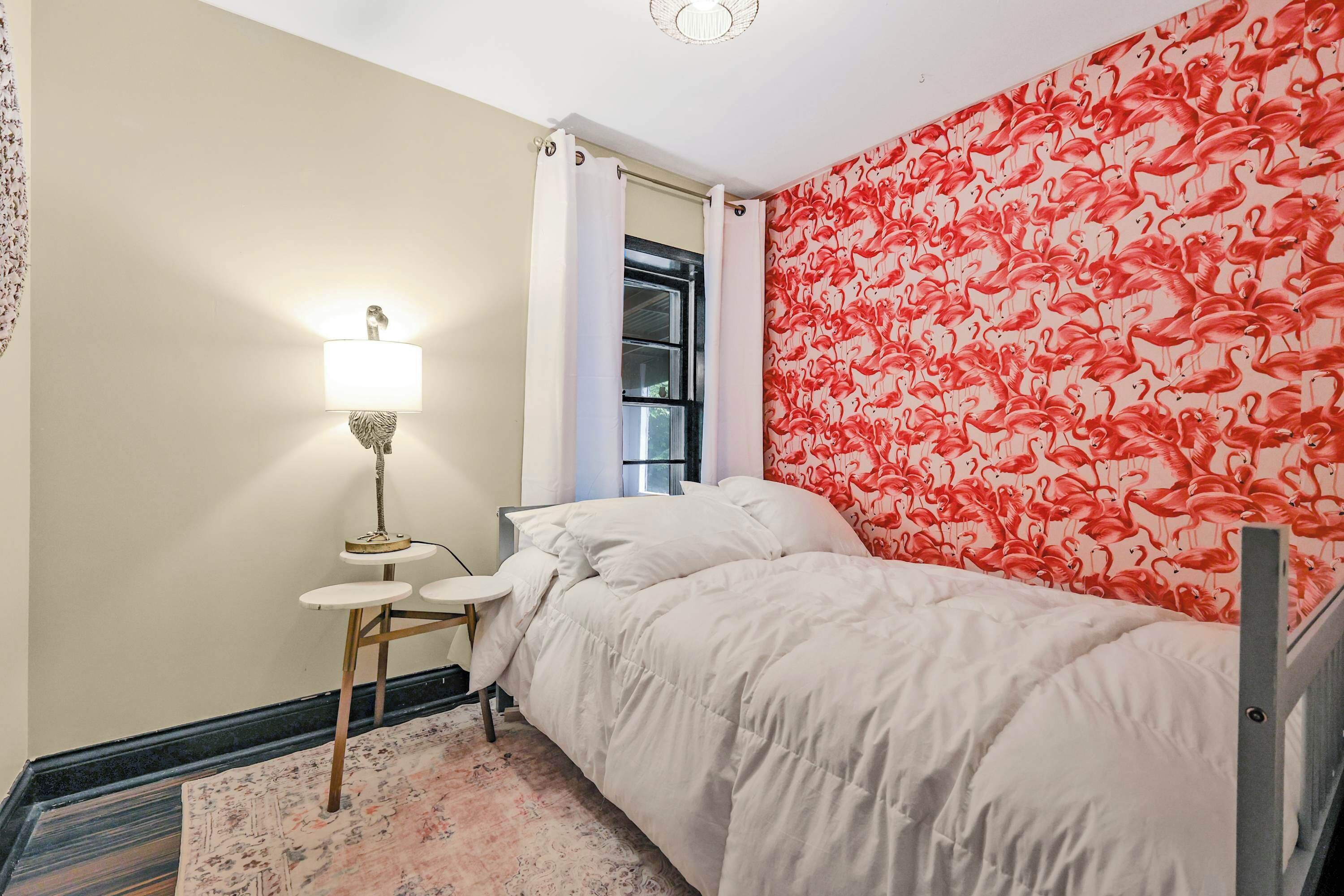 A bedroom with a bed, a nightstand with a lamp, a window with curtains, and a wall with a flamingo pattern.