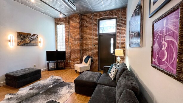A modern living room with exposed brick walls, a large window, and a flat-screen TV. - Writers Round (Nashville)