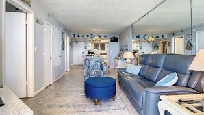 A spacious living room with a blue sofa, a blue ottoman, and a white wicker table. A kitchen is visible in the background. - Oceania Beach Club 805 (New Smyrna Beach)