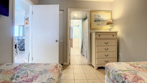A bedroom with two beds, a dresser, a painting, and a door leading to another room. - Oceania Beach Club 805 (New Smyrna Beach)