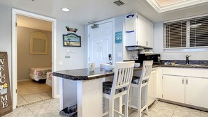 A kitchen with white cabinets, a granite countertop, and a sink. There are two white bar stools and a small island with a sink. - Oceania Beach Club 805 (New Smyrna Beach)