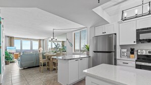 A modern kitchen with white cabinetry, a stainless steel refrigerator, and a dining area with a view of the sea. - Oceania Beach Club 101 (New Smyrna Beach)