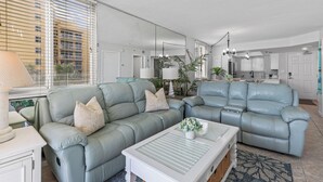 A modern living room with a sectional sofa, a coffee table, and a large window with blinds. - Oceania Beach Club 101 (New Smyrna Beach)