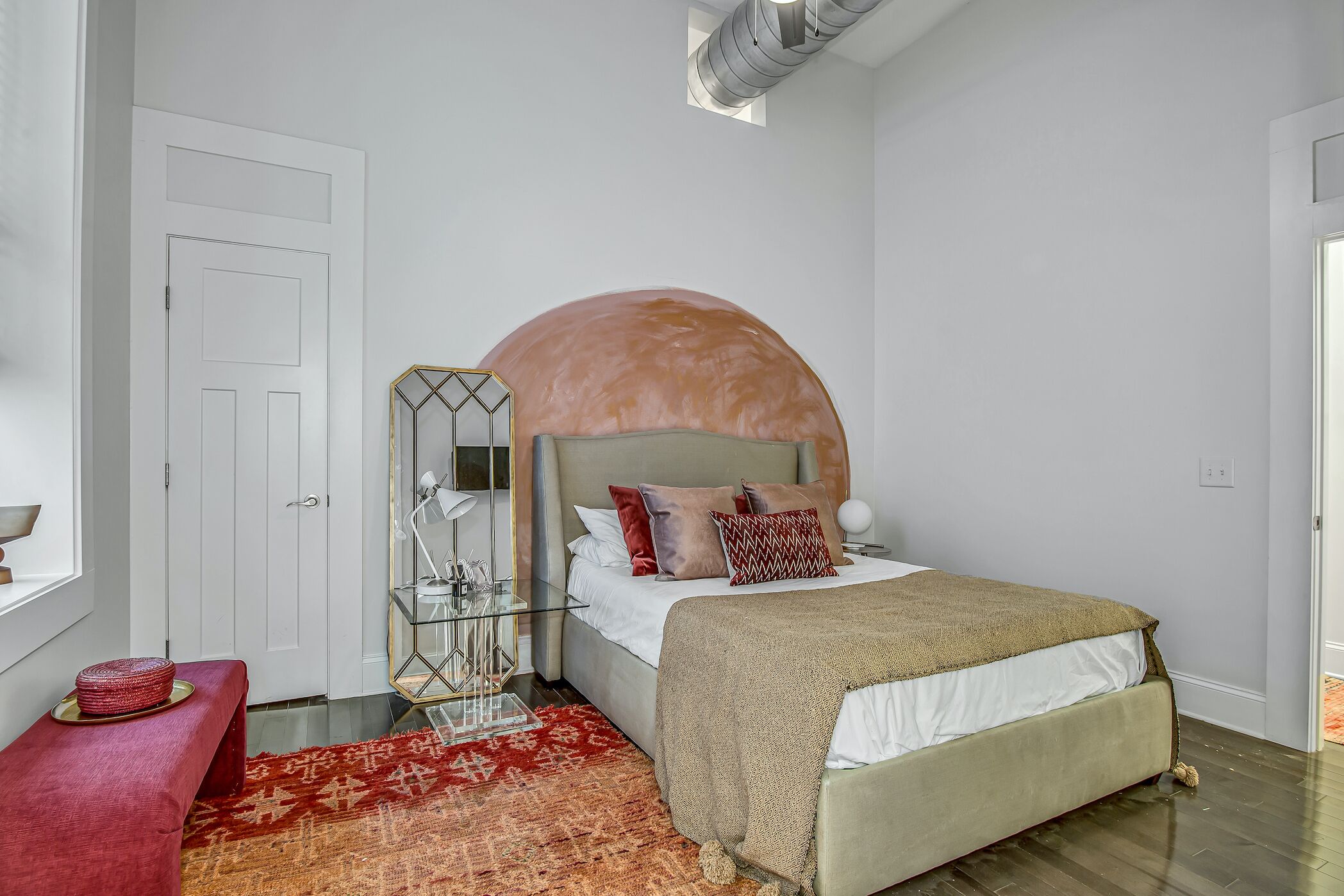 A bedroom with a large bed, a red bench, a glass side table, and a patterned rug.