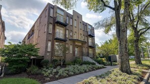 Un immeuble à plusieurs étages, construit en briques, doté de balcons, d’un porche avant couvert et d’un jardin paysager avec des arbres et des arbustes.