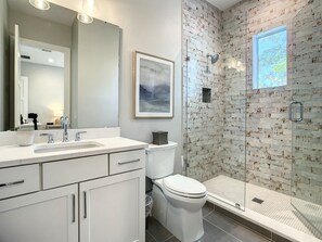 A modern bathroom with a white sink, mirror, and toilet. There is a glass shower enclosure with a view of trees. - Lala at the Beach 512 (New Smyrna Beach)